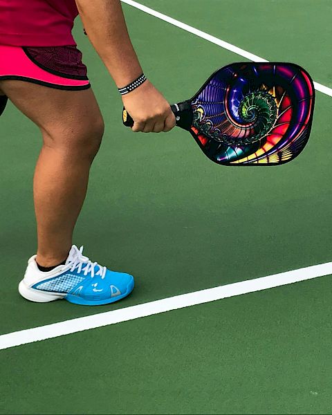 A person in sports attire holding a colorful pickleball paddle on a green court, preparing to play.