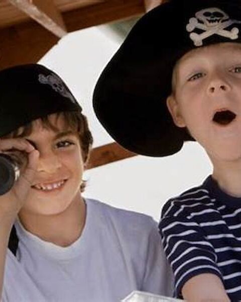 Two children are dressed as pirates, one using a telescope and the other with an open mouth, both wearing pirate hats.