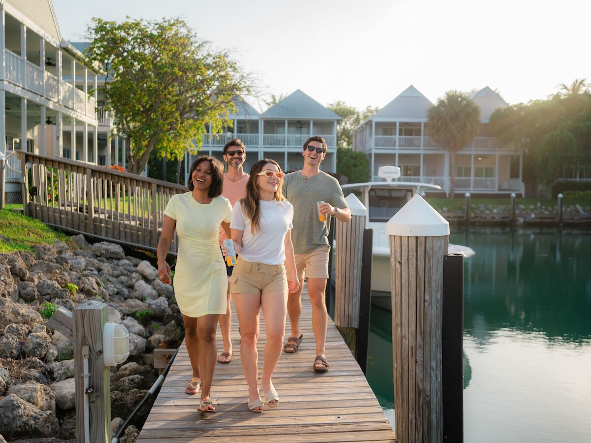 A group of four people walking on a wooden dock by the water, with houses and greenery in the background, in a sunny setting.