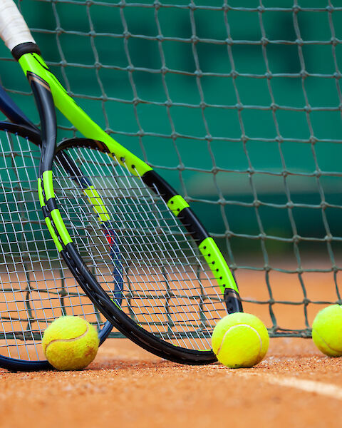 Two tennis rackets are leaning against a net with four tennis balls on a clay court.