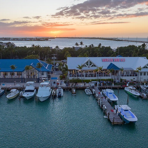 A marina with several boats docked, buildings by the water, and a sunset in the background.