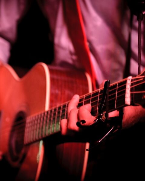 A person is playing an acoustic guitar with a capo on the neck, wearing a shirt with rolled-up sleeves, under low red lighting.
