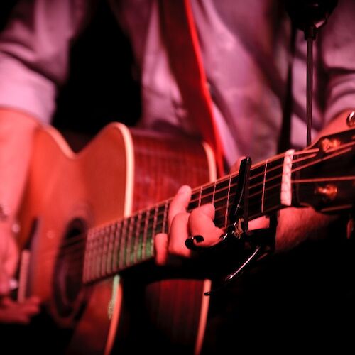 A person is playing an acoustic guitar with a capo on the neck, wearing a shirt with rolled-up sleeves, under low red lighting.