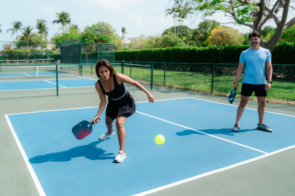 Two people playing pickleball on an outdoor court, with one player hitting the ball while their partner watches.