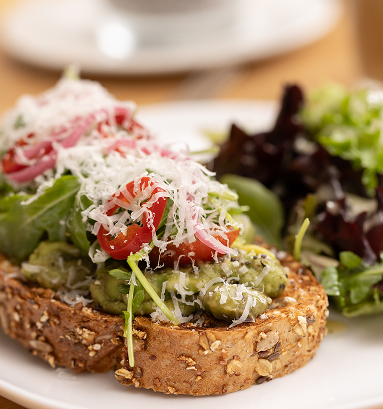 Avocado toast topped with cherry tomatoes, shredded cheese, and microgreens on seeded bread, served with a side salad.