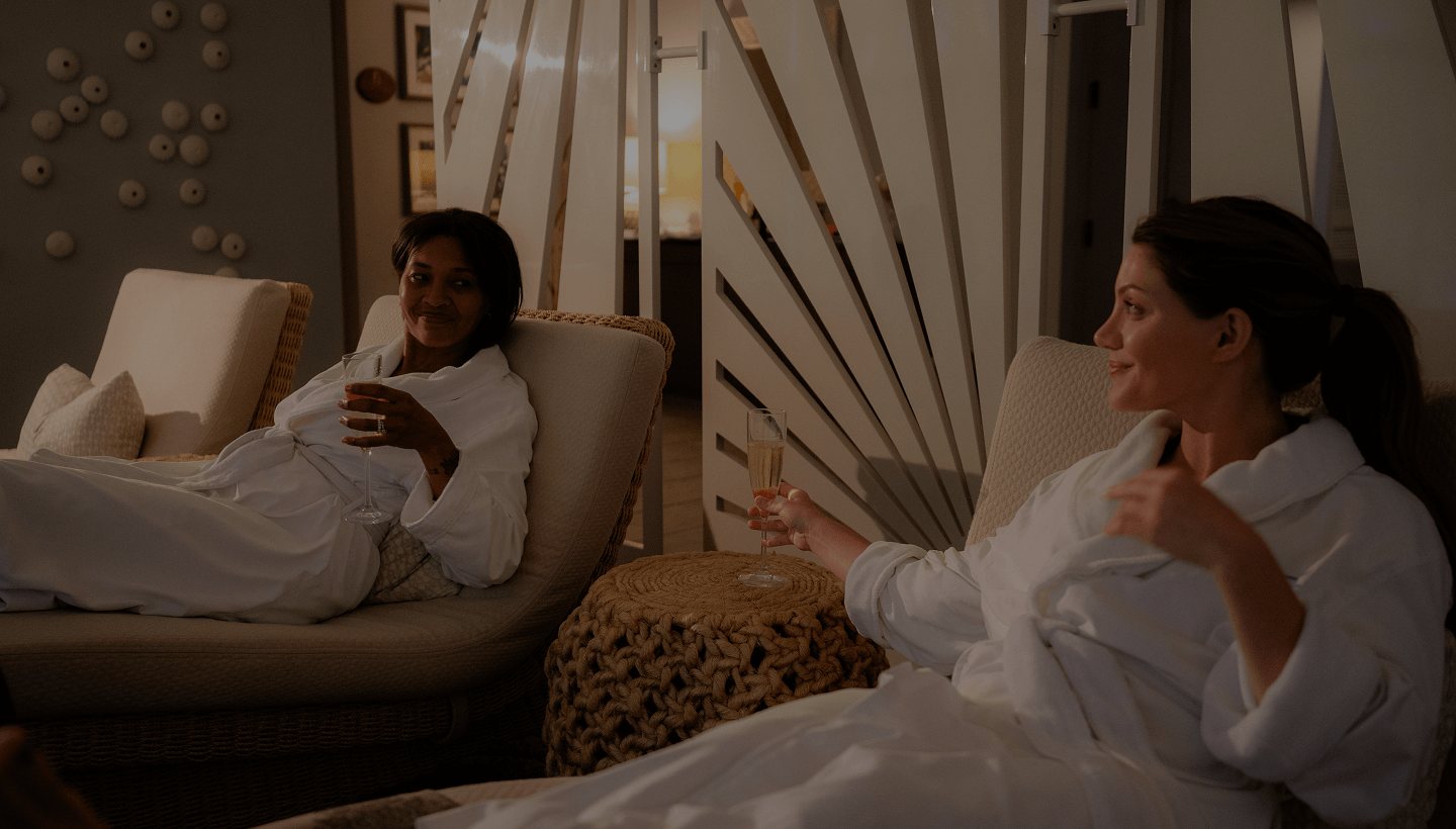Two women in spa robes relaxing in lounge chairs, chatting and holding champagne glasses in a calm, softly lit spa setting.