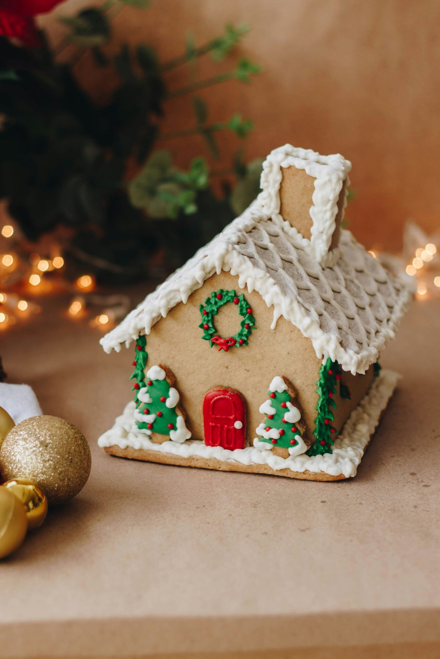 Gingerbread house decorated with icing, wreath, and Christmas trees, set beside gold ornaments and warm holiday lights.