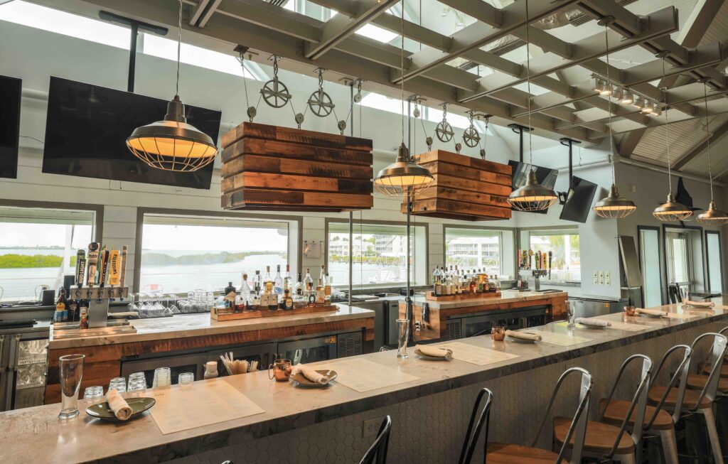 Modern waterfront bar with wood accents, hanging lights, stocked shelves, and counter seating overlooking the water.