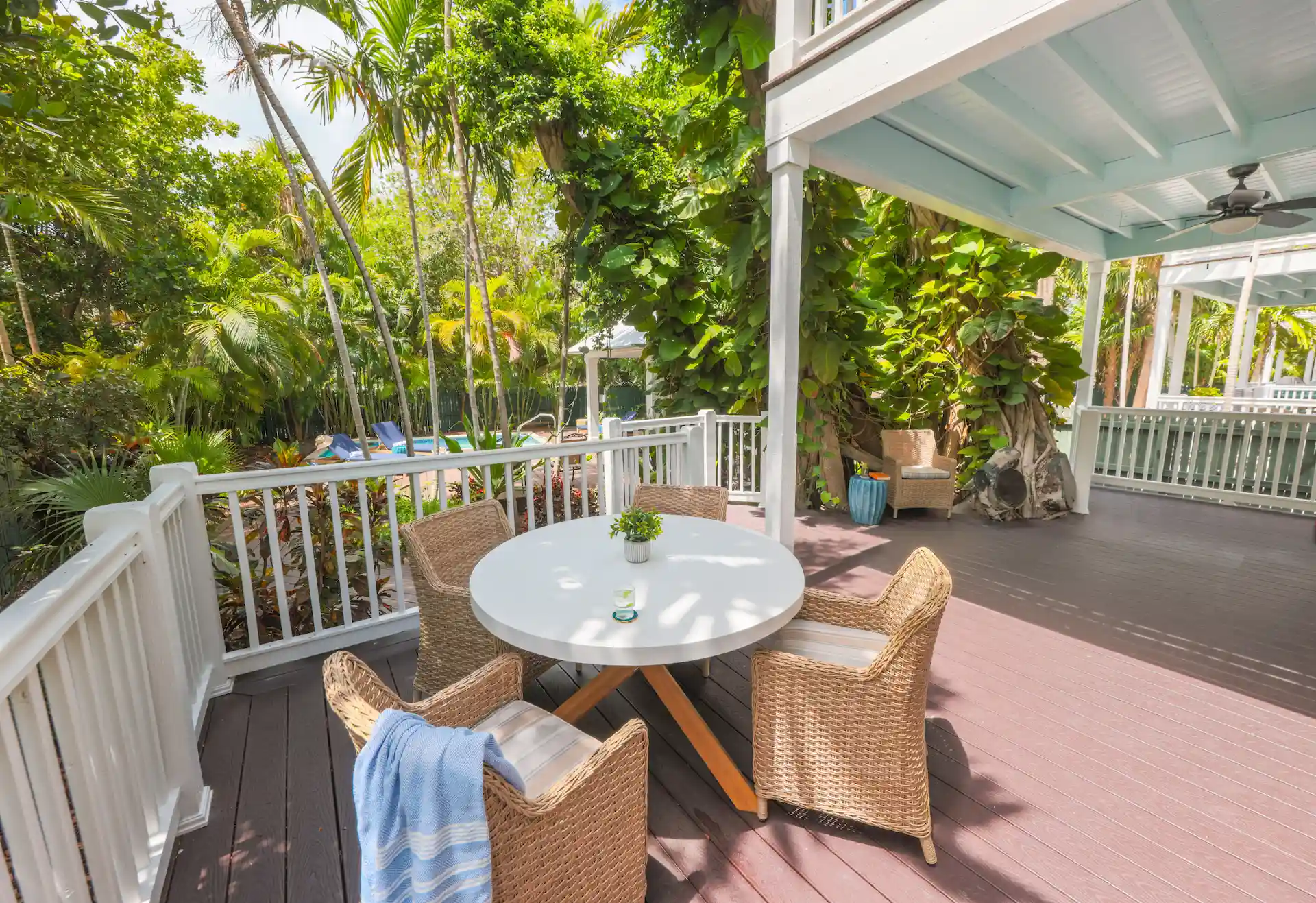 Tropical patio with white table, wicker chairs, striped towel, and pool view surrounded by palm trees.