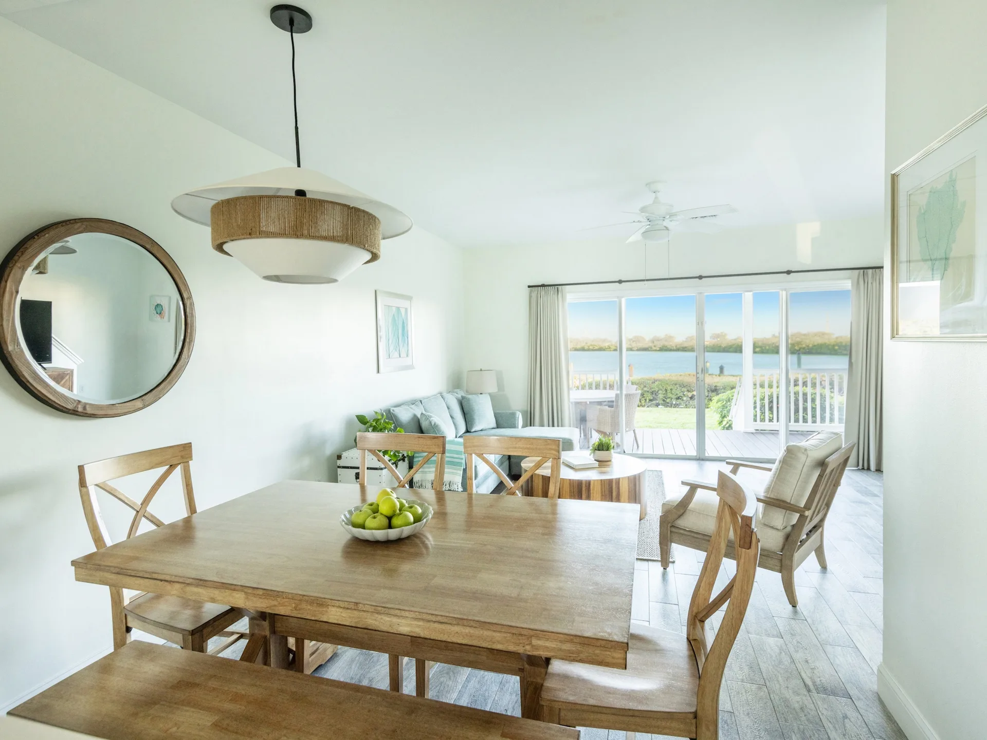 Open-plan dining and living area with wooden table, light blue sofa, and water view through glass doors.