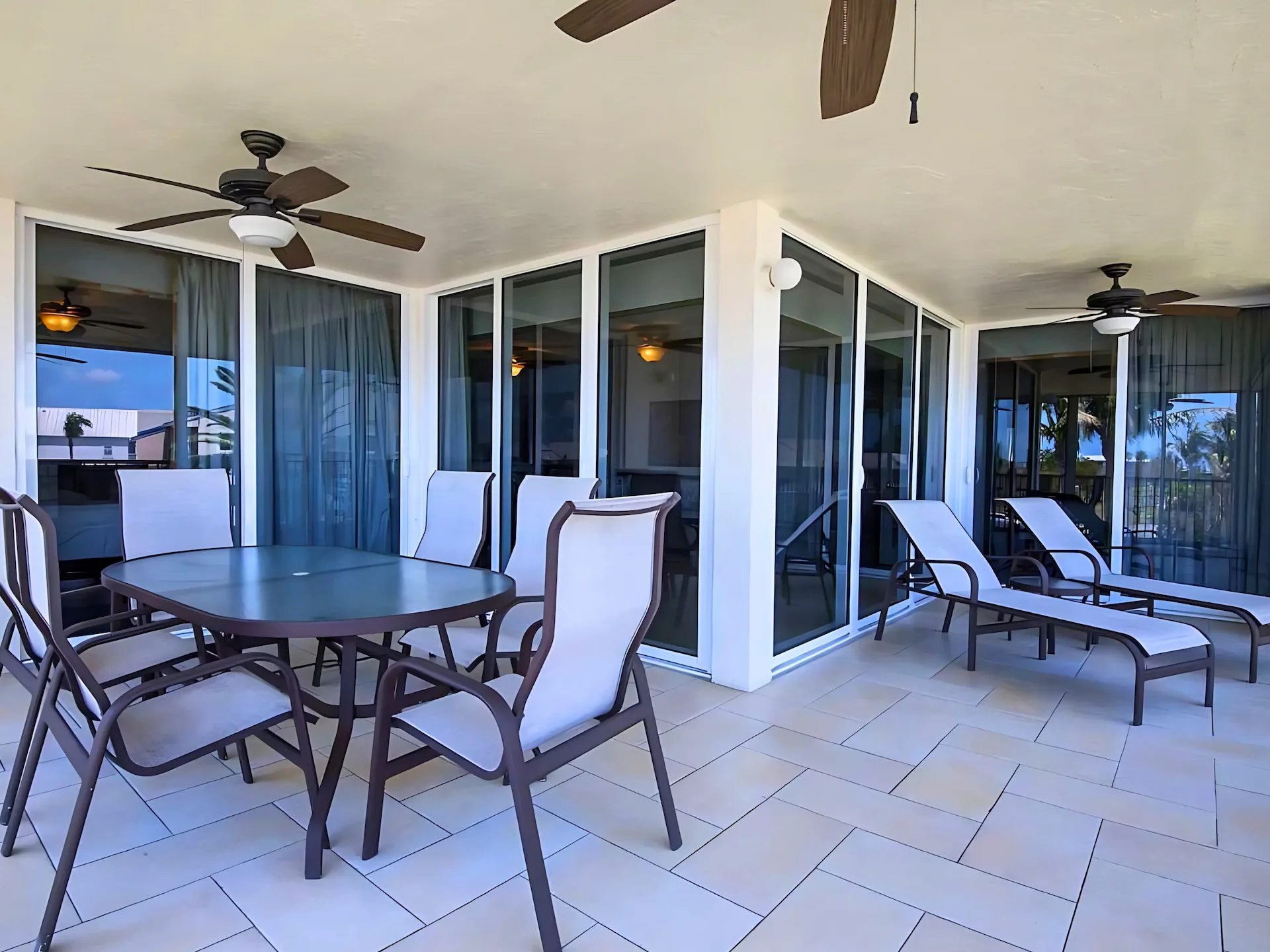 Covered outdoor patio with cushioned dining and lounge chairs, ceiling fans, and view of palm trees through glass doors.