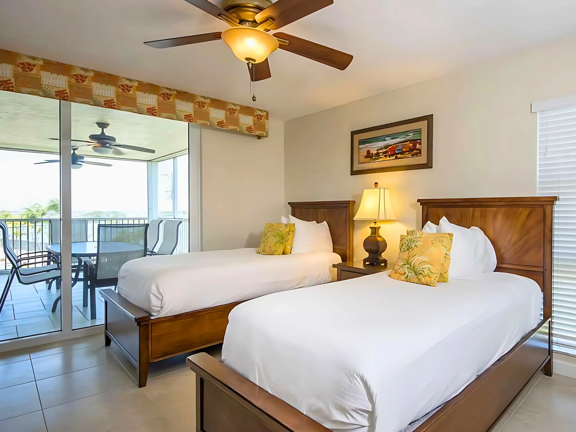 Bedroom with two single beds, yellow floral pillows, coastal artwork, and balcony access with outdoor seating.