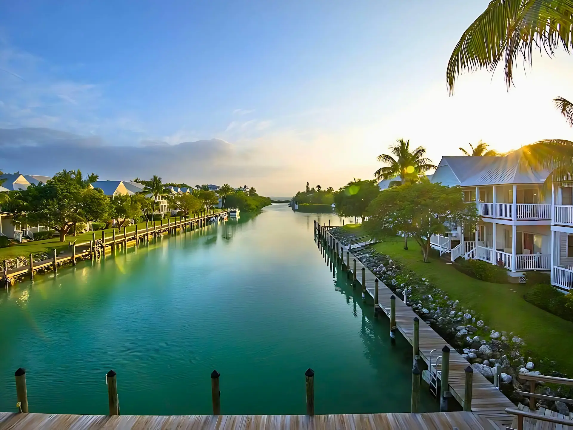 Waterfront homes with white exteriors and docks lining a calm canal at sunrise, surrounded by palm trees and greenery.