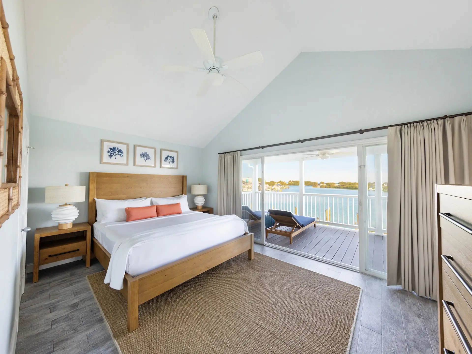 Airy coastal bedroom with wooden bed, orange pillows, coral artwork, and balcony view of water and landscape.
