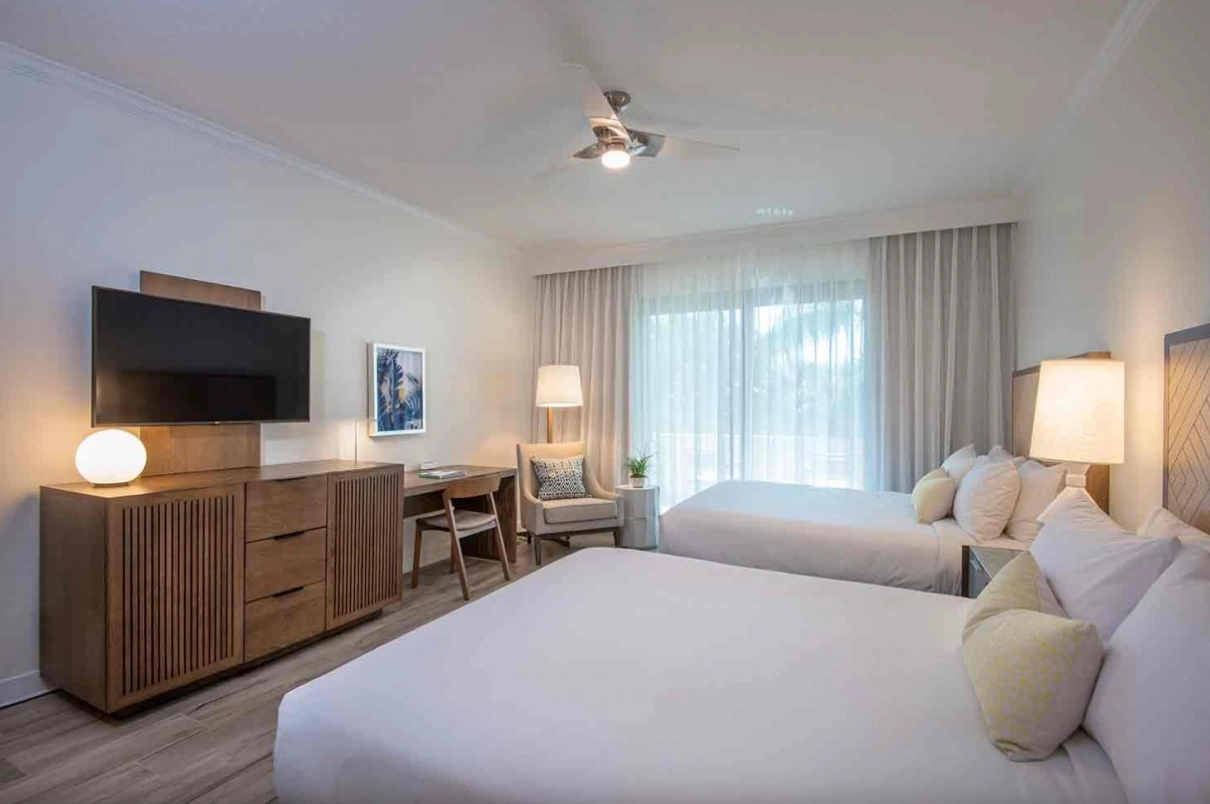 Bright hotel room with two queen beds, wooden furnishings, desk, TV, and balcony doors letting in natural light.