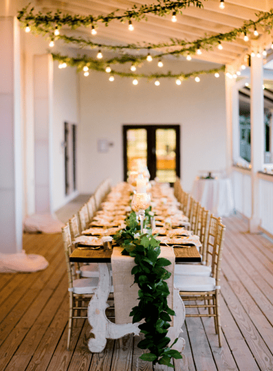 Long decorated dining table with greenery and string lights set on a covered outdoor patio for an elegant event.