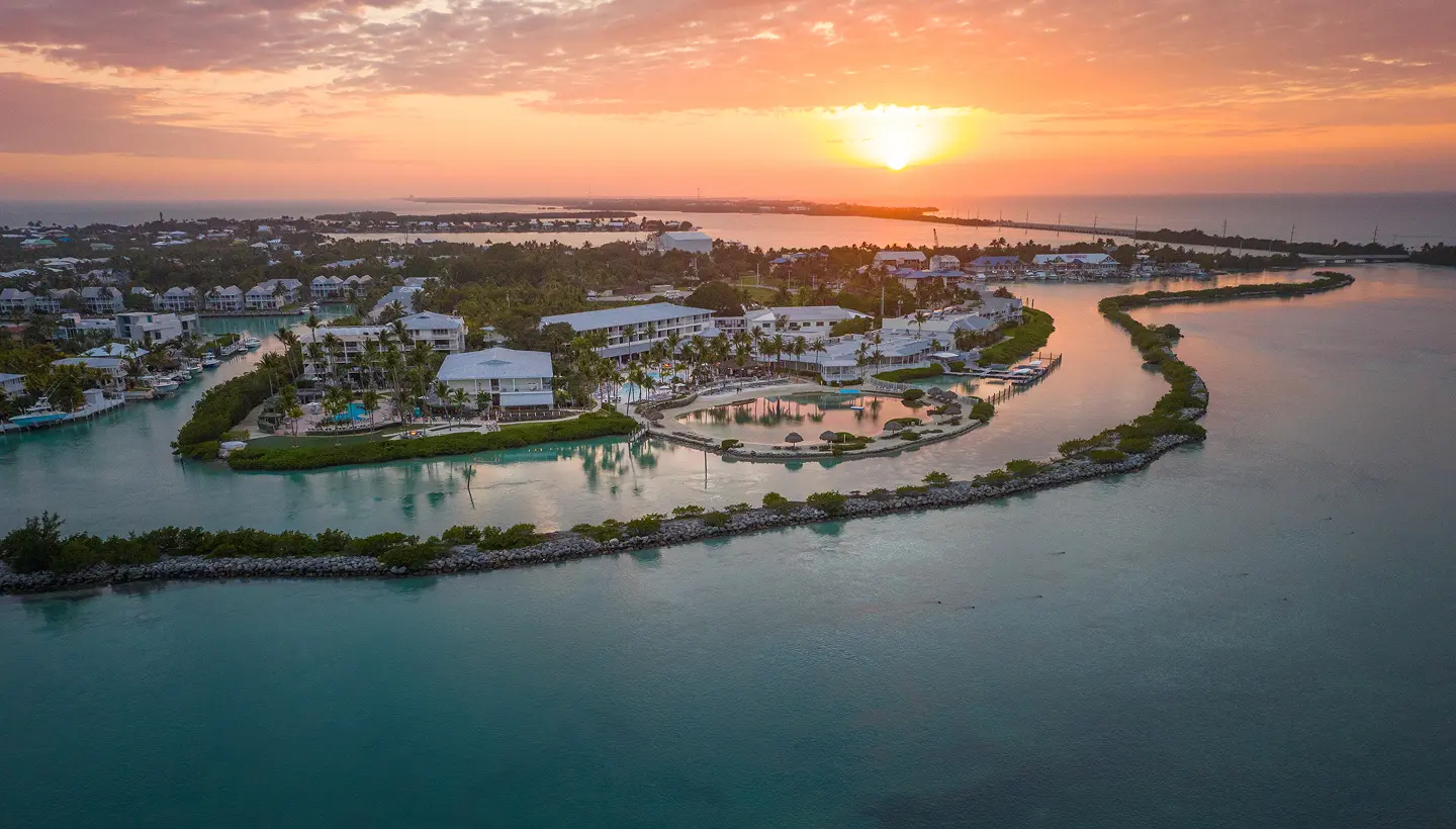 Aerial view of a tropical island resort at sunset, with white buildings, palm trees, and calm turquoise water.
