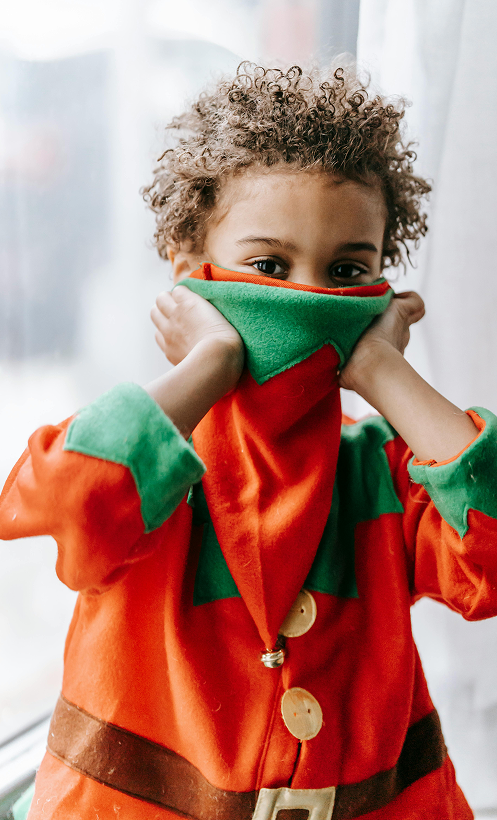 Child in an elf costume covering their face with the collar, looking playfully at the camera indoors.