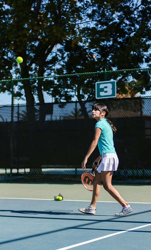 Young girl playing tennis on an outdoor court, watching the ball while holding a racket.