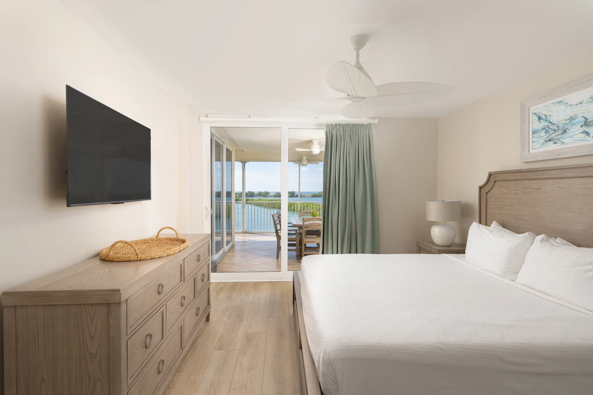 Bedroom with white linens, wooden furniture, wall-mounted TV, and balcony overlooking water and greenery.