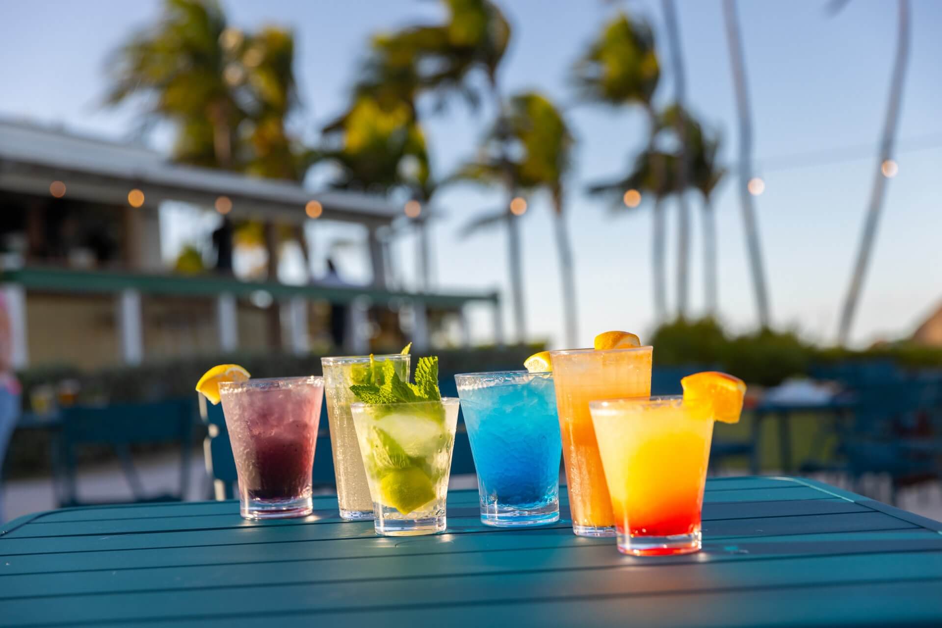 Colorful tropical cocktails lined up on an outdoor table with palm trees and a beachside bar in the background.