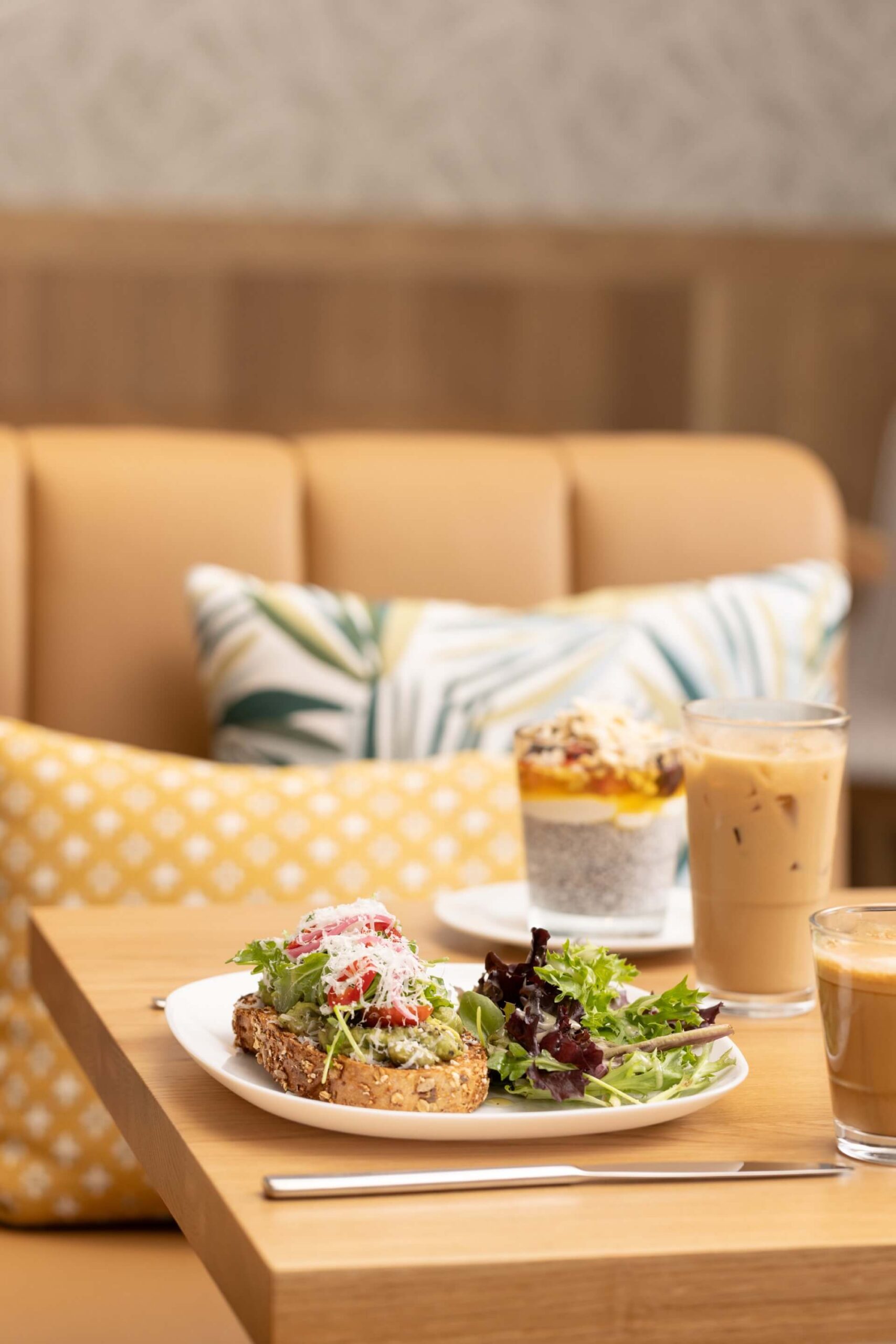 Cafe table with avocado toast, side salad, iced coffee, and parfait in a cozy, modern seating area.