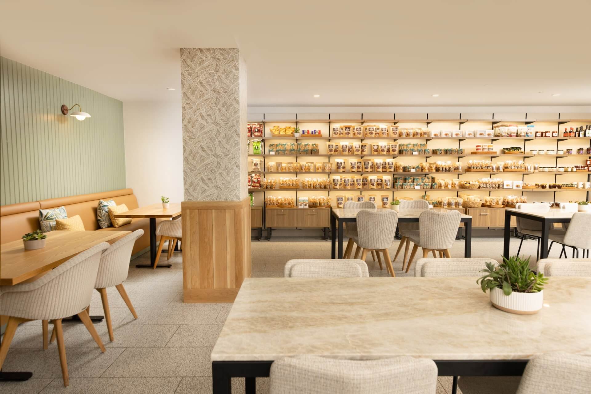 Modern café seating area with wooden tables, upholstered chairs, and shelves of packaged snacks along the back wall.