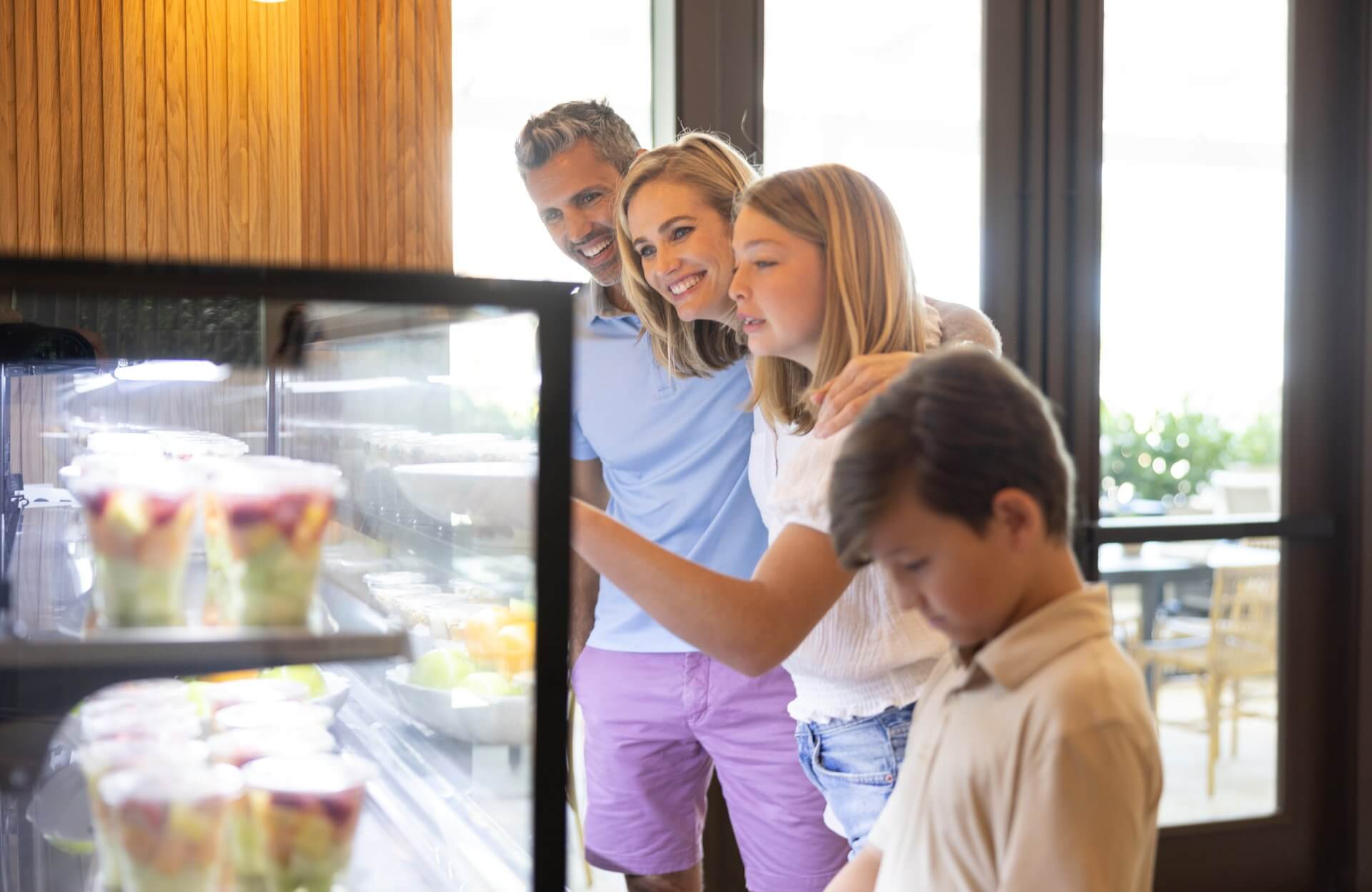 Family browsing a café display case with fresh desserts and fruit cups, smiling together in a bright indoor setting.