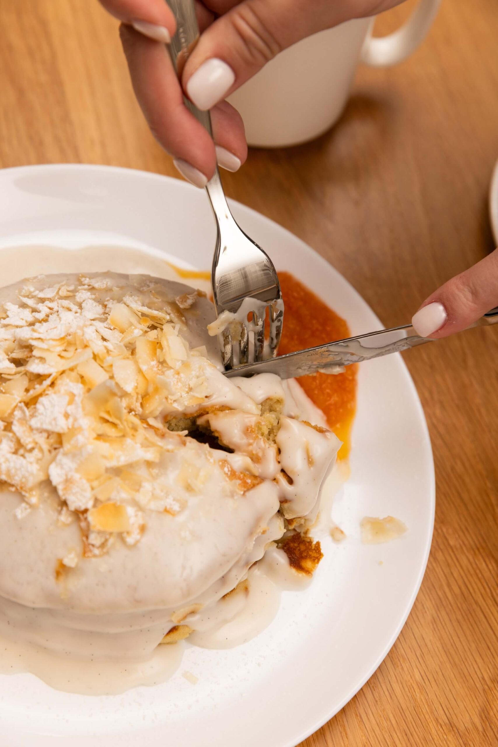 Hands cutting into a glazed pastry topped with sliced almonds on a white plate.