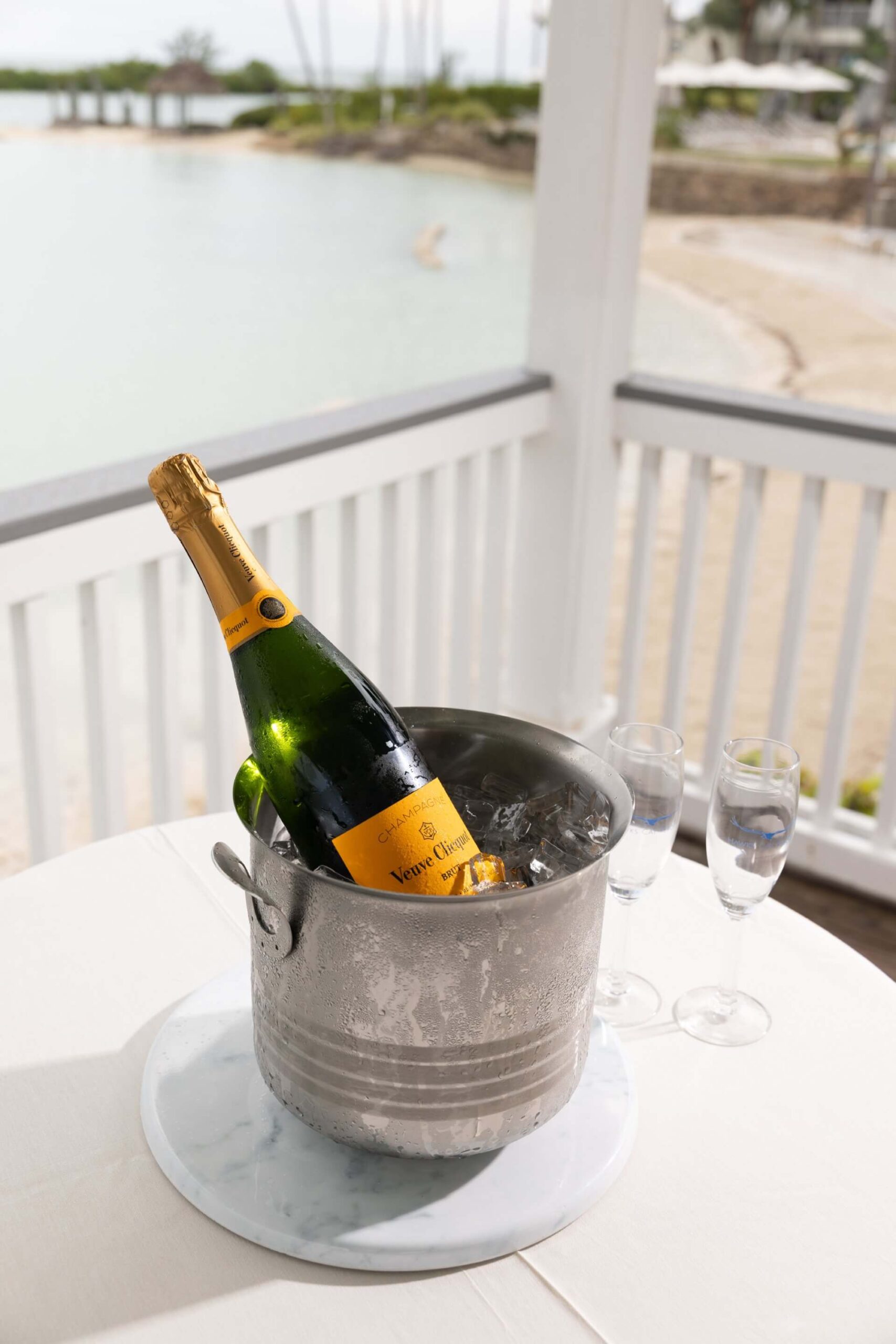 Champagne bottle chilling in an ice bucket with two flutes on a table overlooking a beachside lagoon.