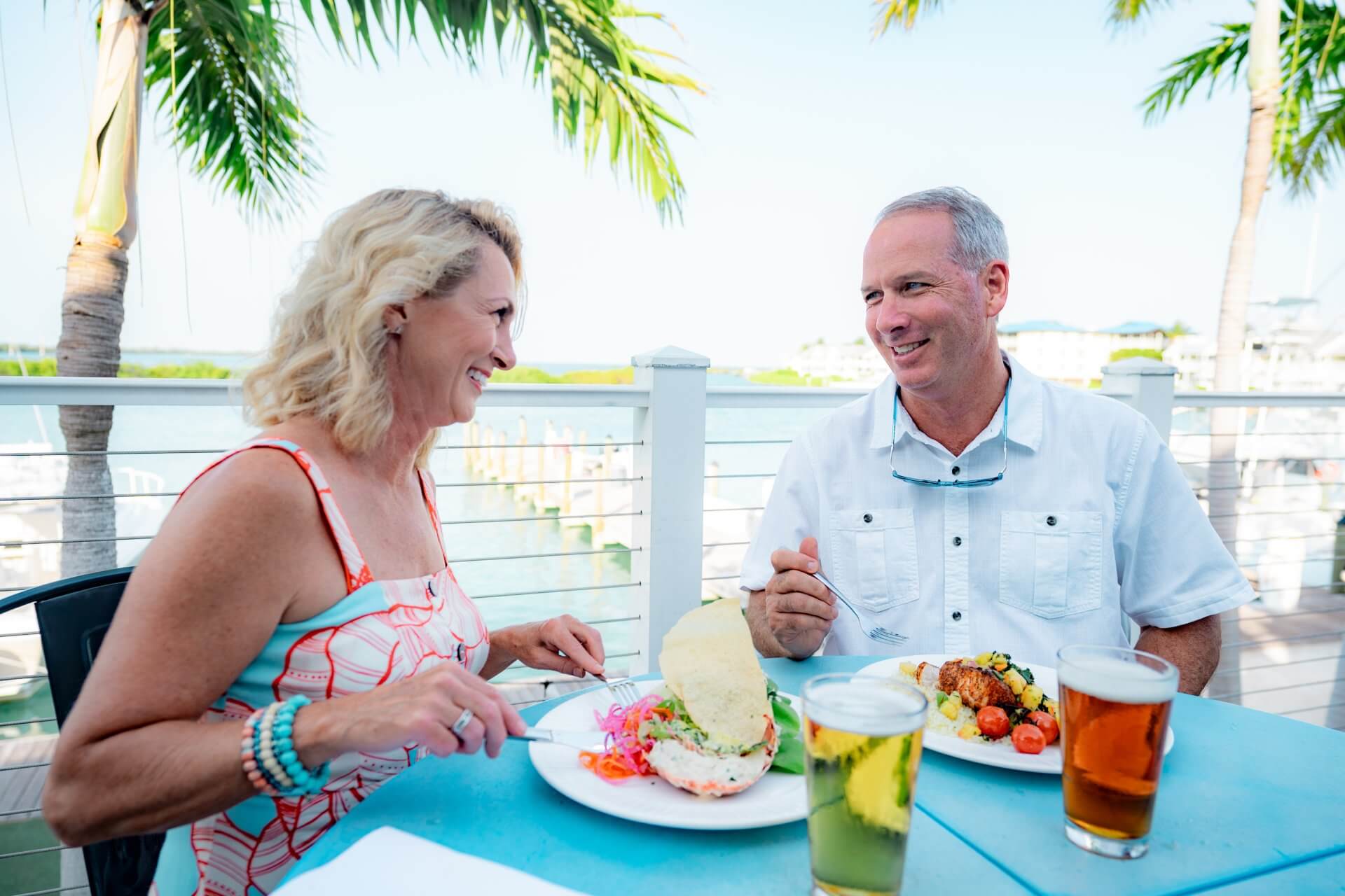 Couple enjoying a waterfront meal, smiling at each other over plates of food and drinks on a sunny patio with palm trees.