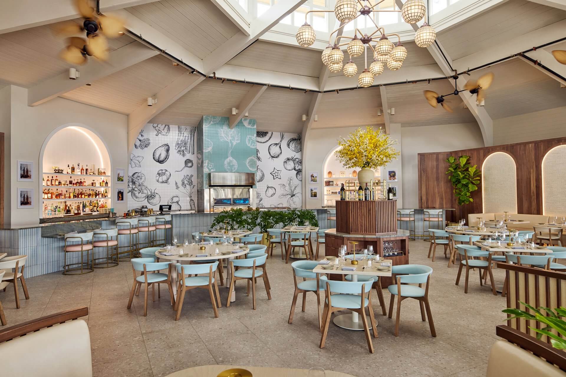 Bright, modern restaurant with vaulted ceilings, chandeliers, a central bar, and pastel dining chairs arranged around round tables.