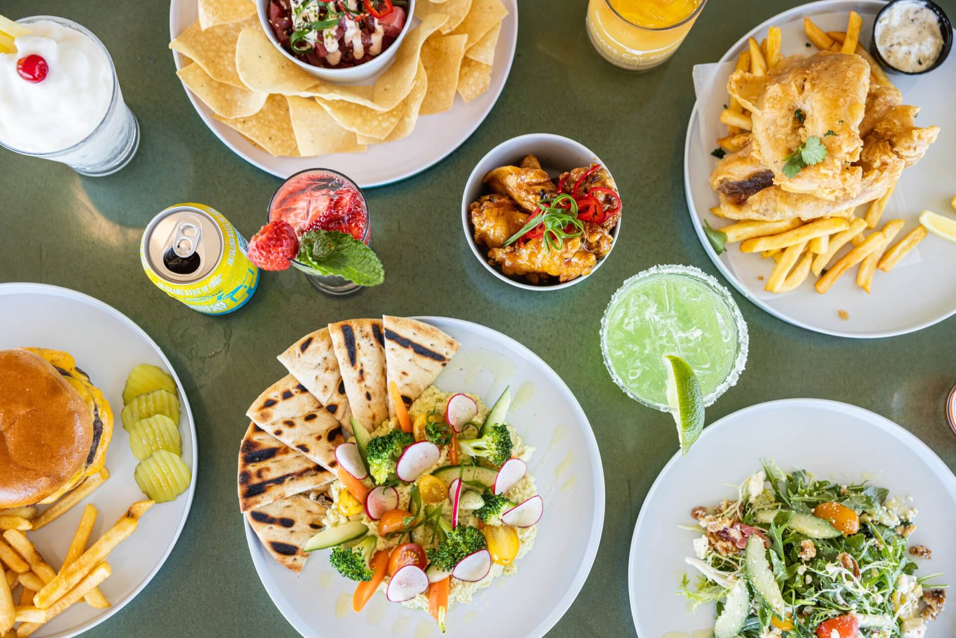 Overhead view of a restaurant table with burgers, fish and chips, salads, chips and dips, flatbread, and colorful cocktails.