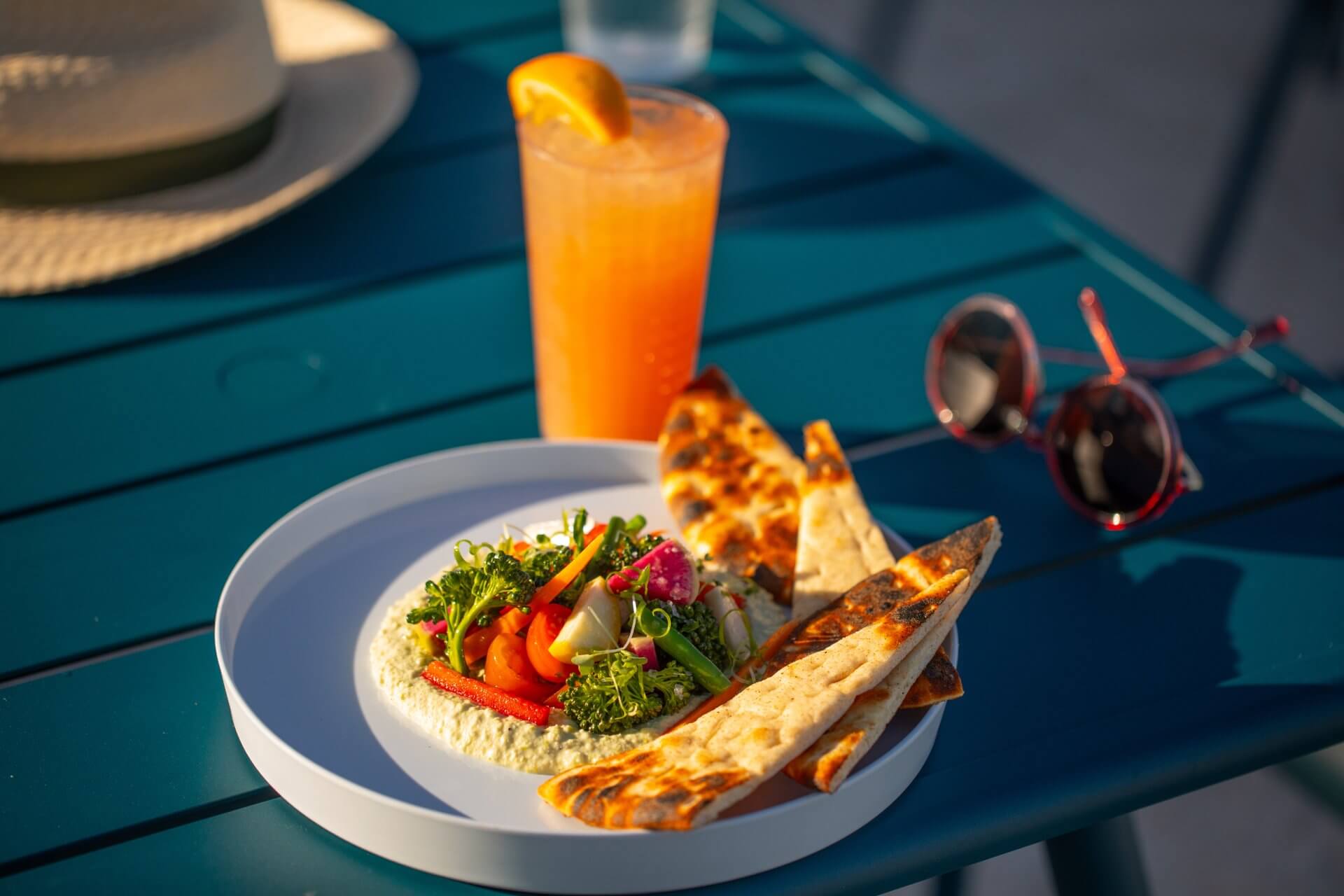 Plated Mediterranean-style dish with grilled pita, fresh vegetables, and an orange cocktail on a sunlit outdoor table.