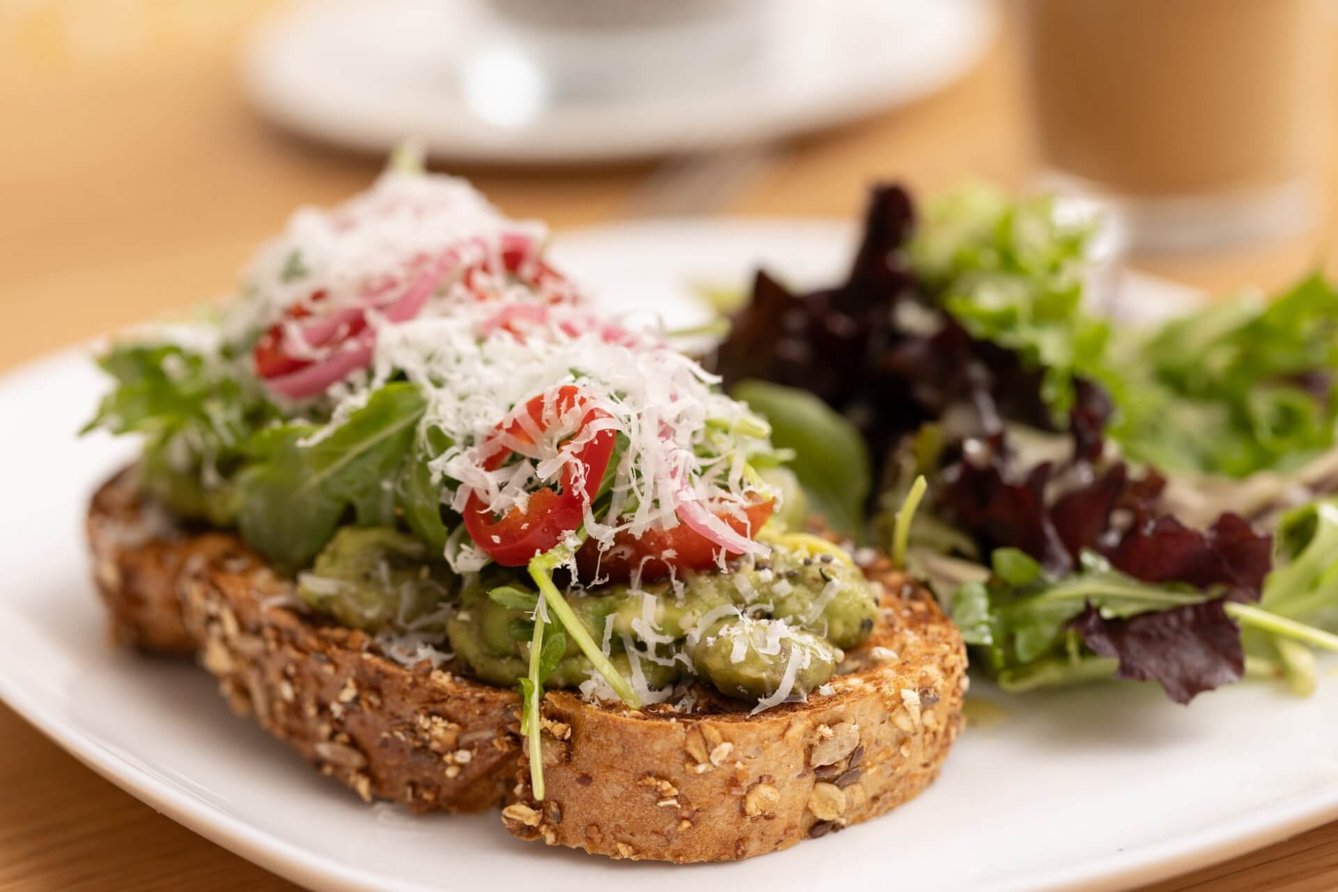 Avocado toast topped with cherry tomatoes, shaved cheese, and greens, served with a fresh side salad on a white plate.