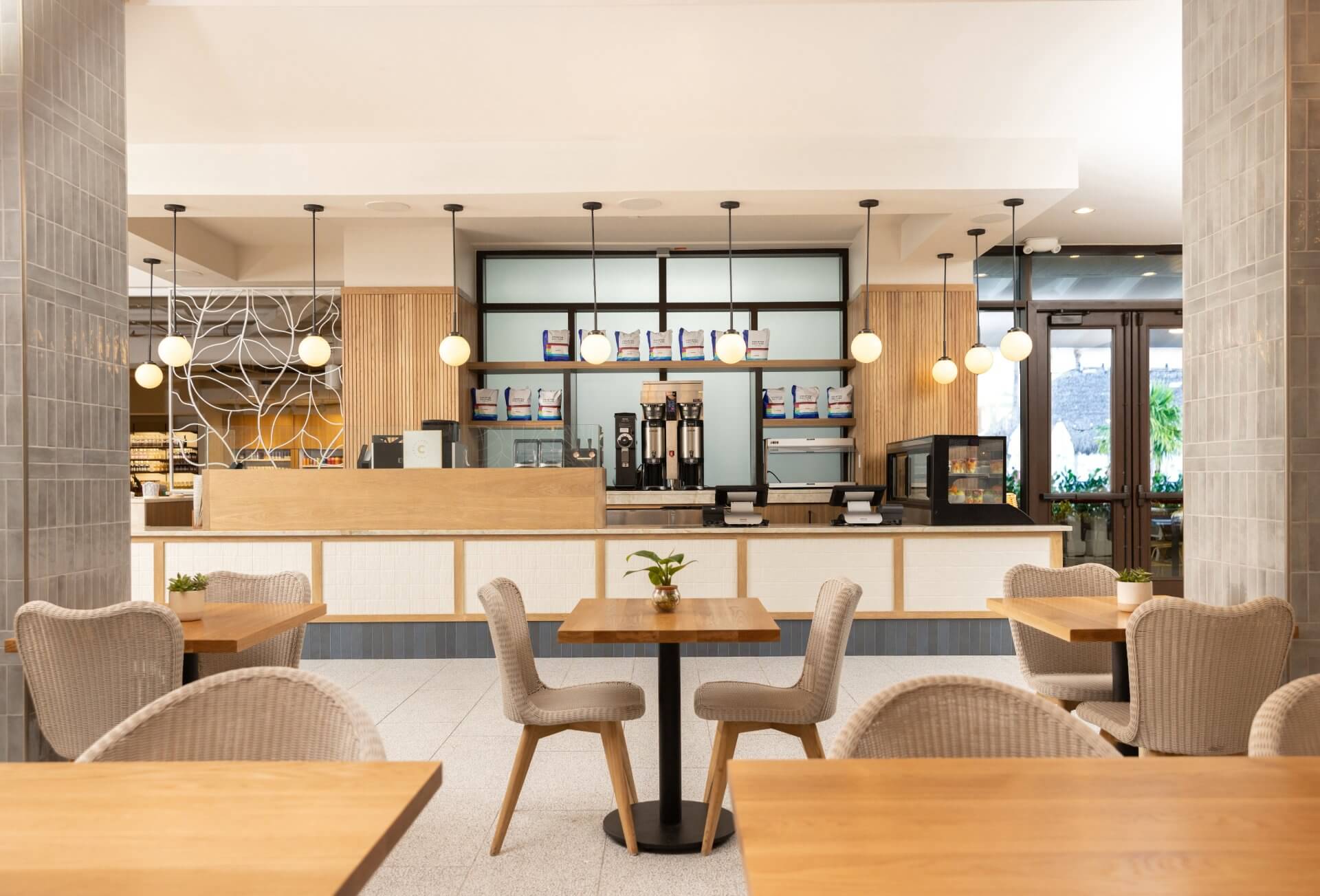 Modern café interior with wooden tables, woven chairs, pendant lights, and a coffee counter with espresso machines.