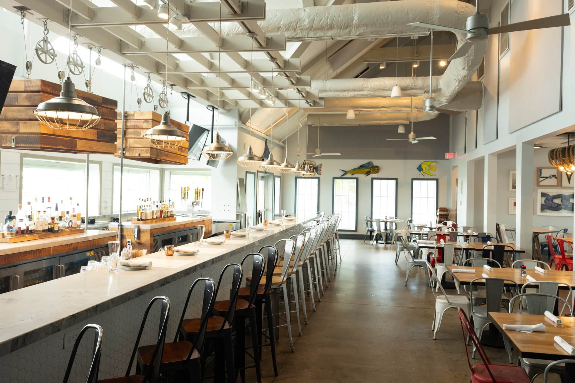 Bright coastal restaurant interior with a long bar, high stools, hanging lights, and dining tables in an open space.