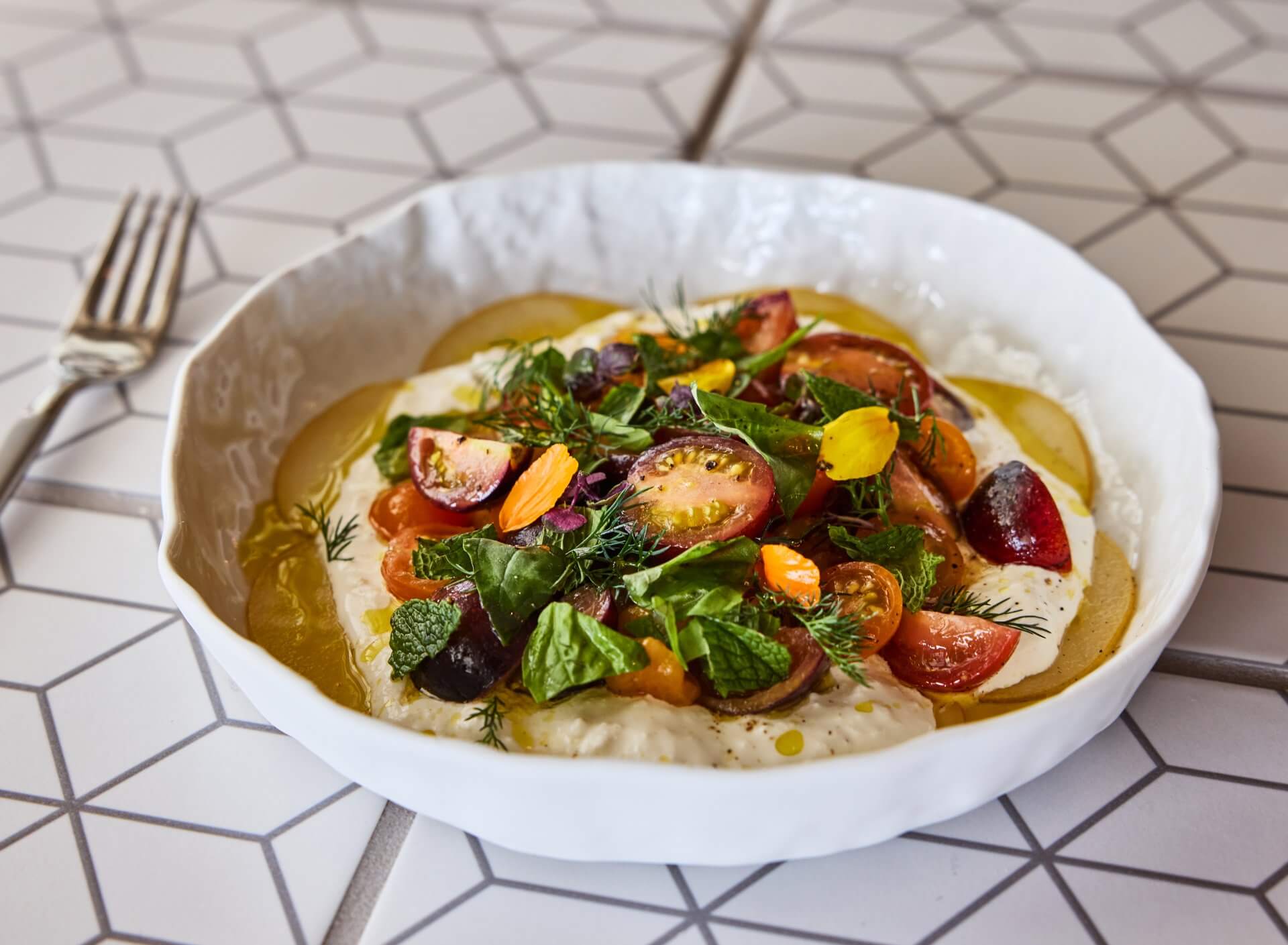 Creamy hummus topped with heirloom tomatoes, fresh herbs, olive oil, and edible flowers in a white bowl.