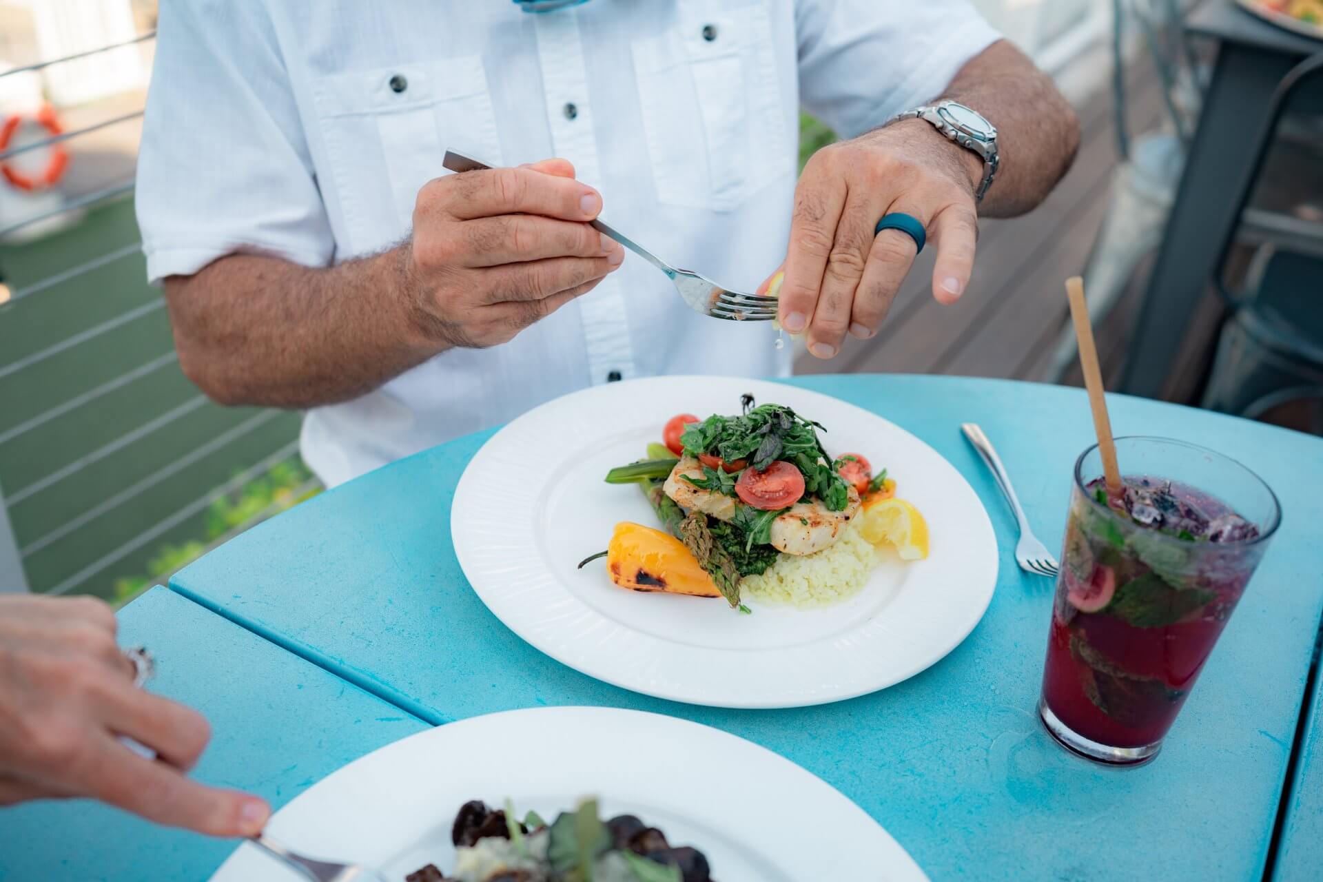 Person eating a plated seafood dish with vegetables at an outdoor table, with a cocktail beside the plate.