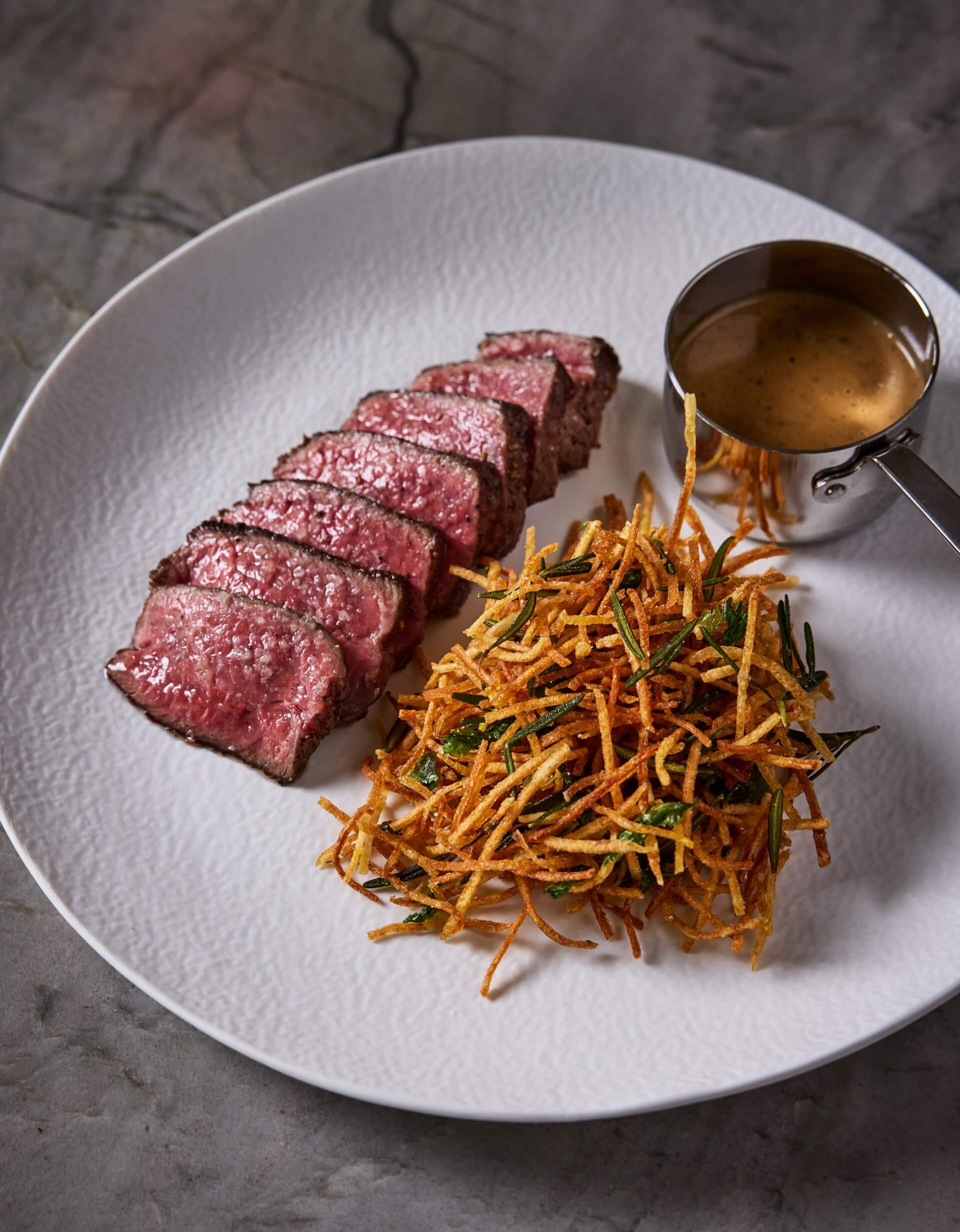 Sliced medium-rare steak served with crispy shoestring potatoes and a small cup of brown sauce on a white plate.