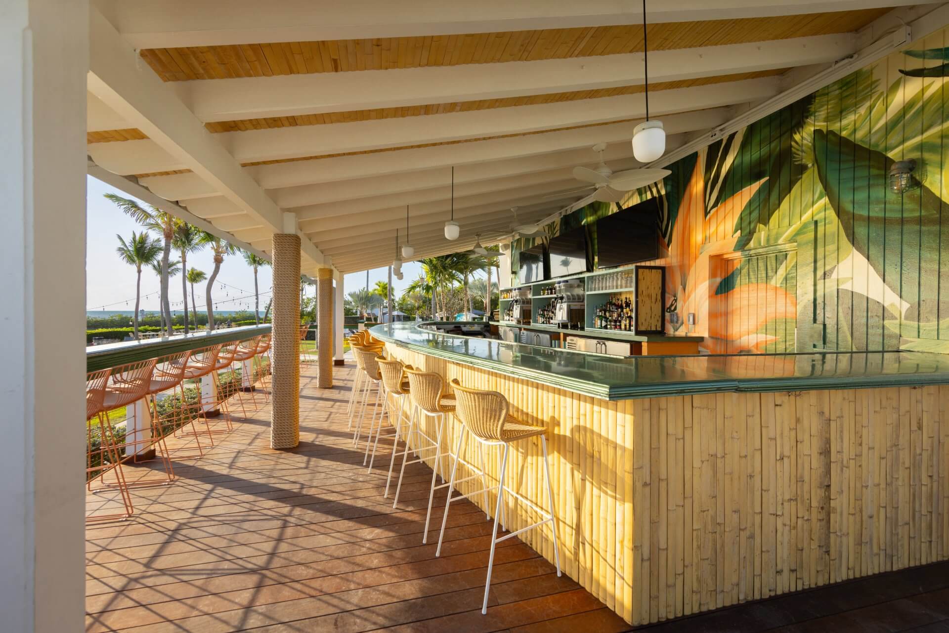 Open-air tropical bar with bamboo counter, wicker stools, mural wall, and palm-lined views in warm afternoon light.