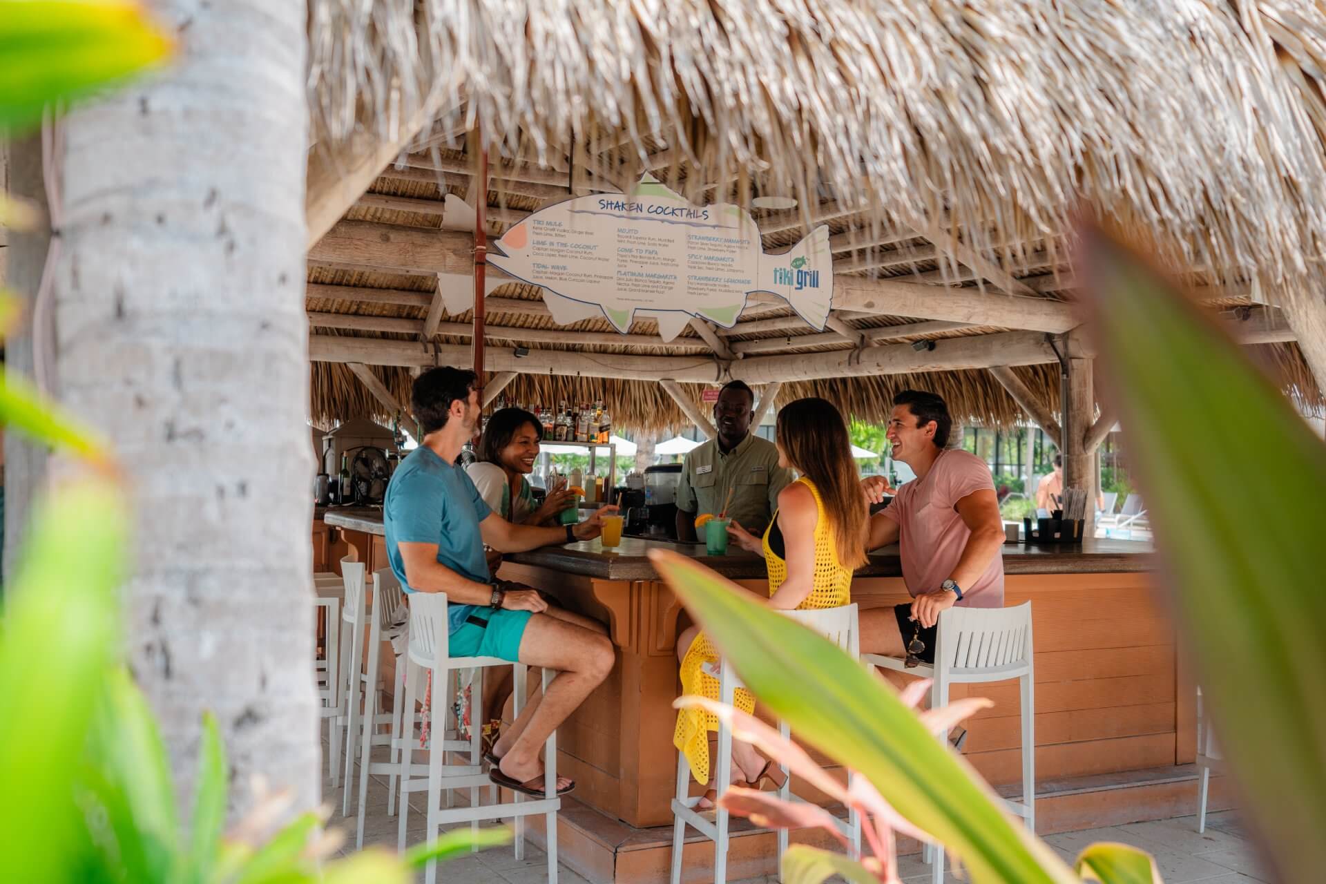 Group of friends enjoying drinks at a thatched tiki bar, chatting with a bartender in a tropical resort setting.