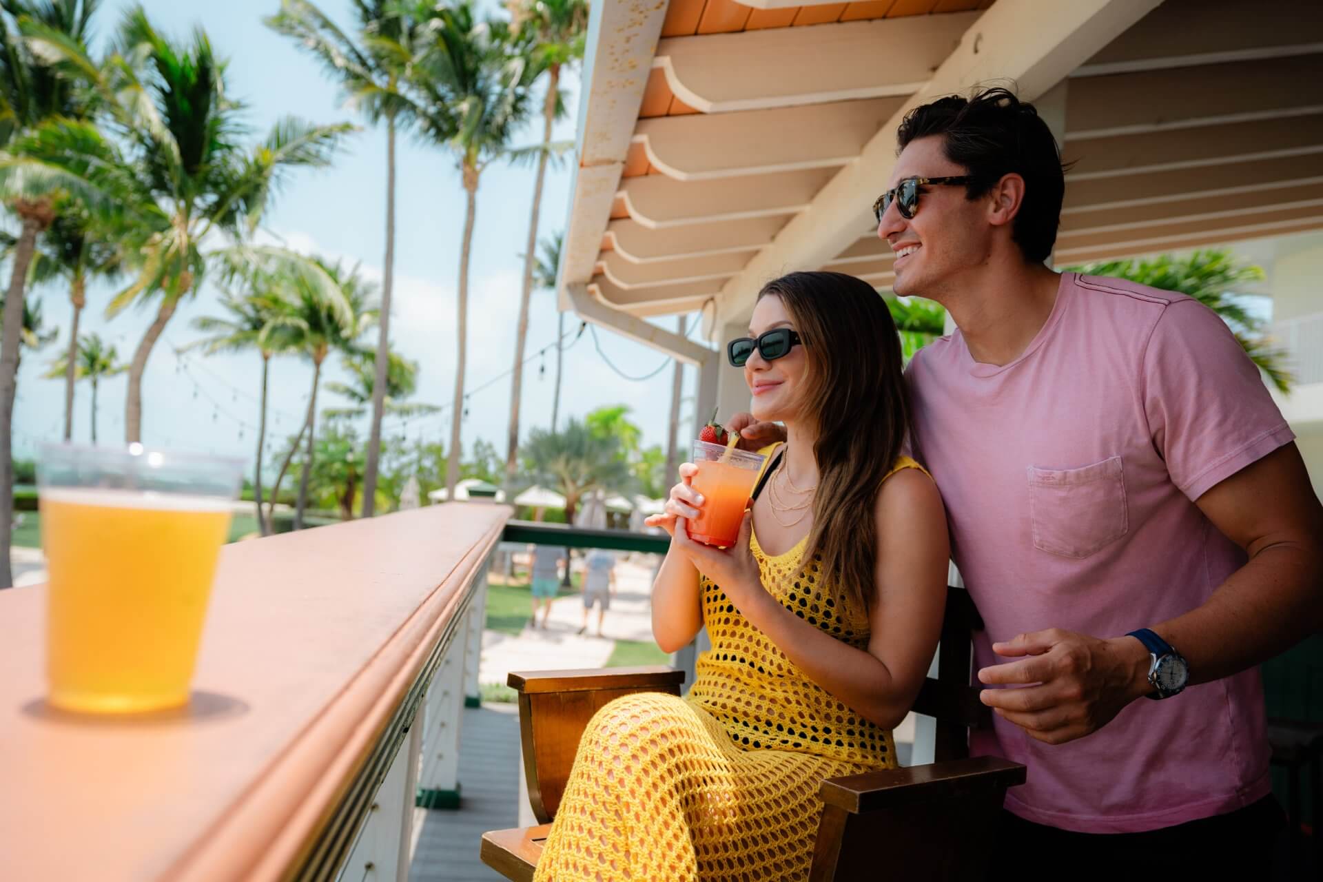 Couple enjoying tropical drinks on a sunny resort balcony with palm trees and ocean breeze.