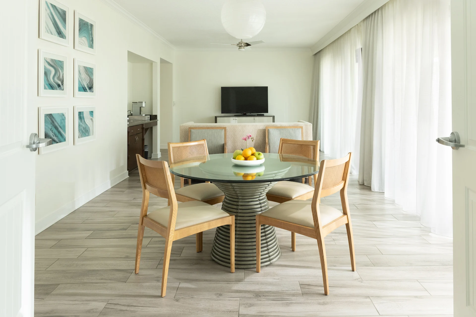 Bright dining space with glass table, fruit bowl, and cozy living area with sofa and TV.