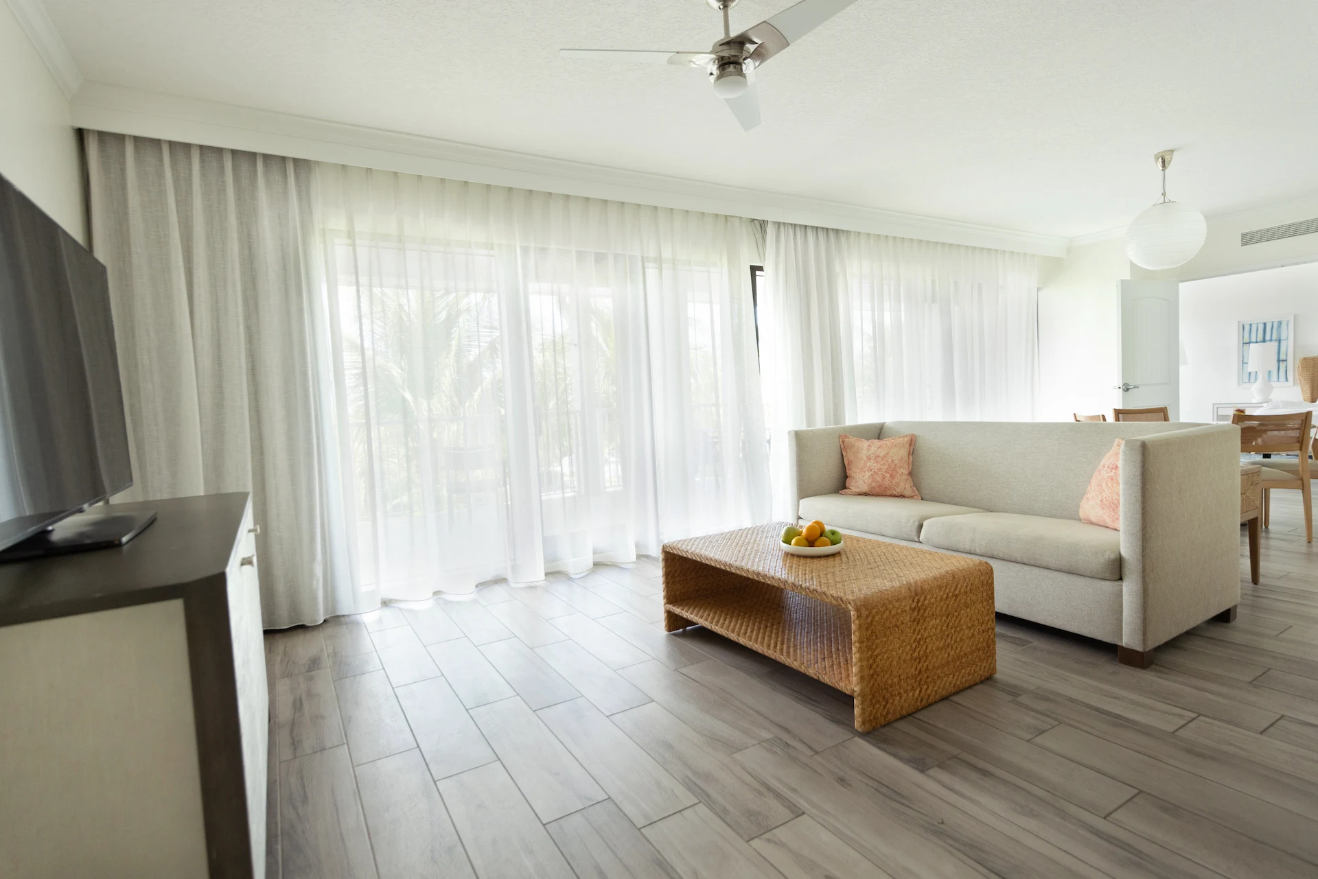 Airy living room with light sofa, rattan coffee table, and large windows with sheer curtains.