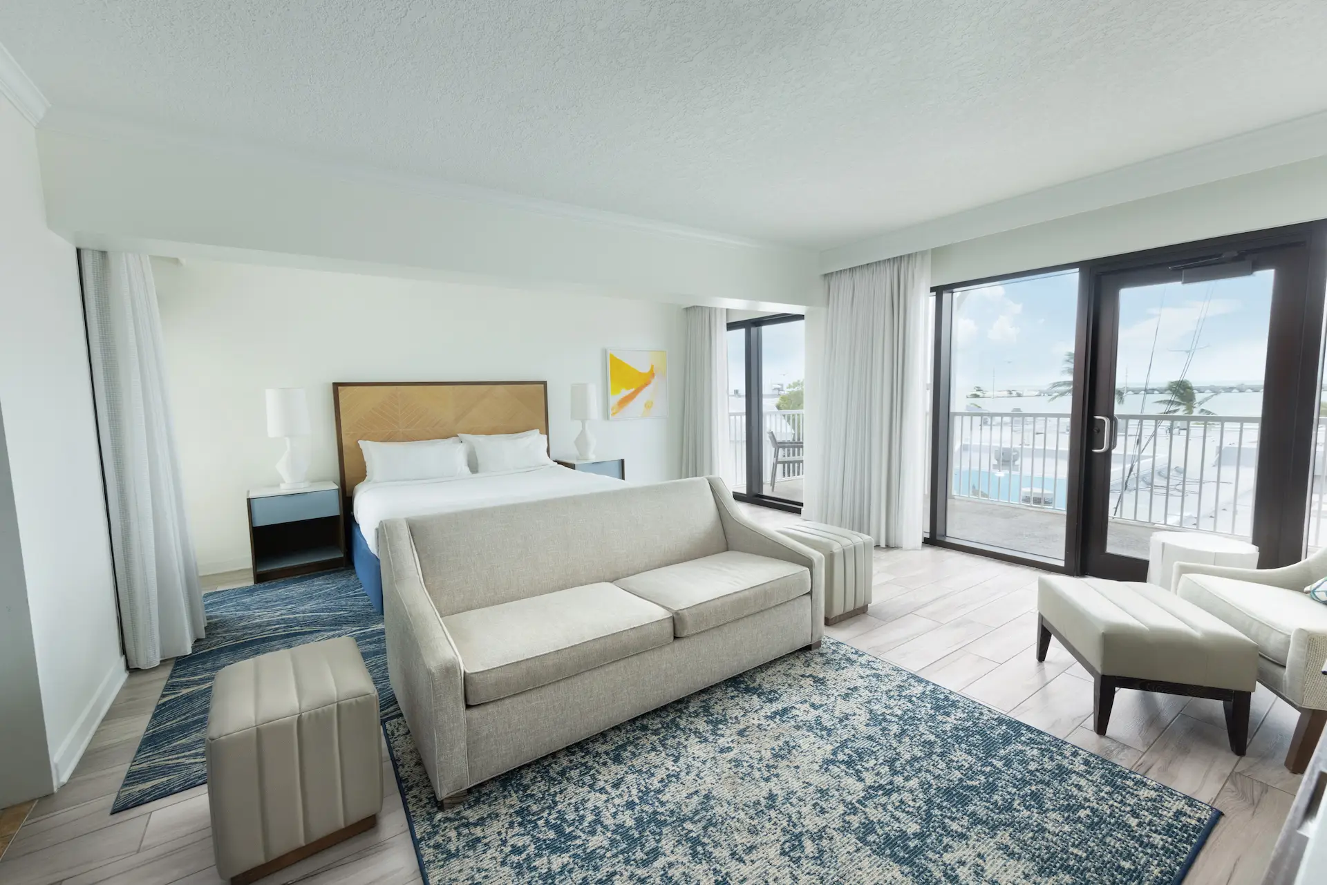 Inviting room with white bed, loveseat, and balcony overlooking a marina in natural light.