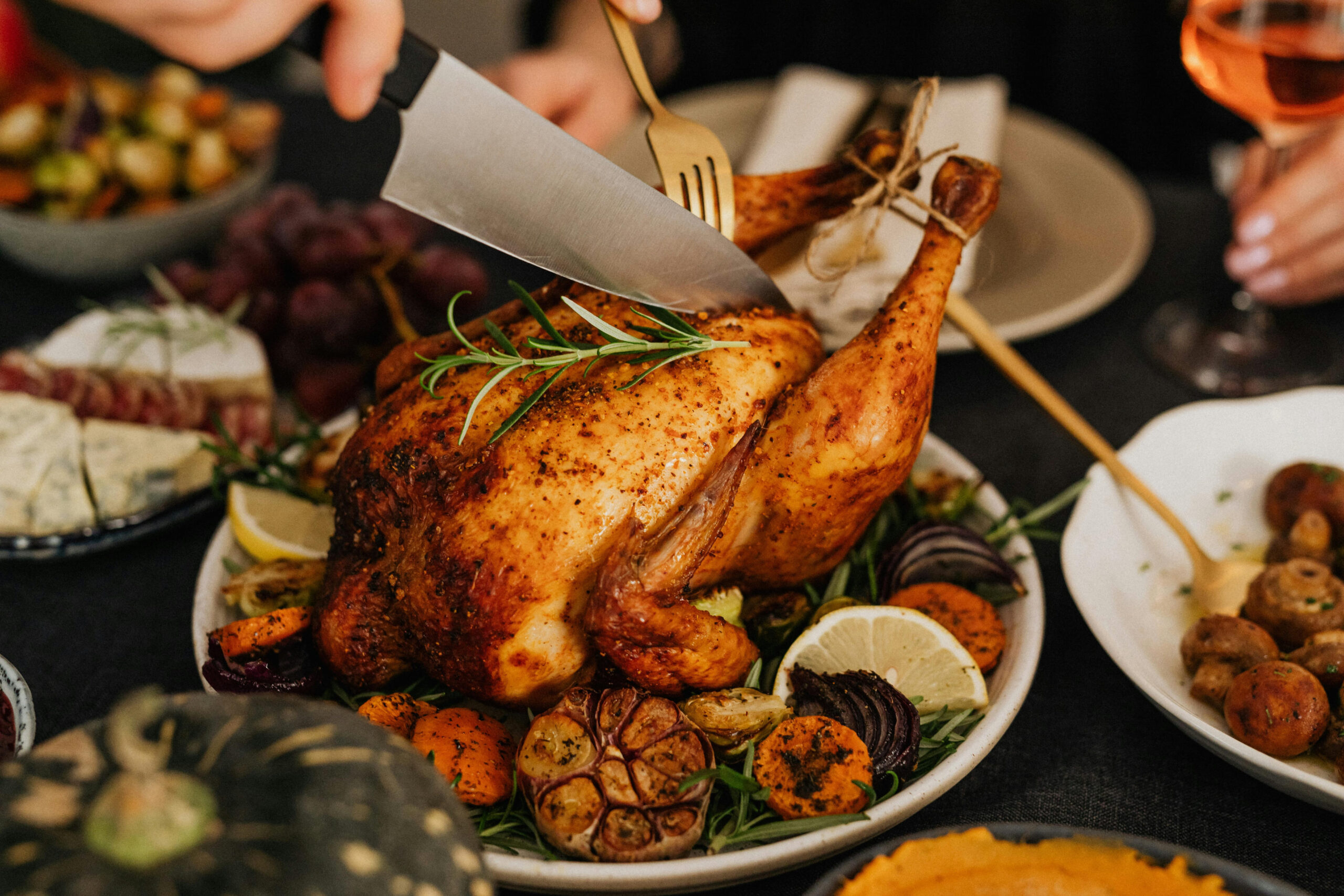 Roasted whole chicken garnished with rosemary and lemon, surrounded by vegetables on a dinner table as someone carves it.