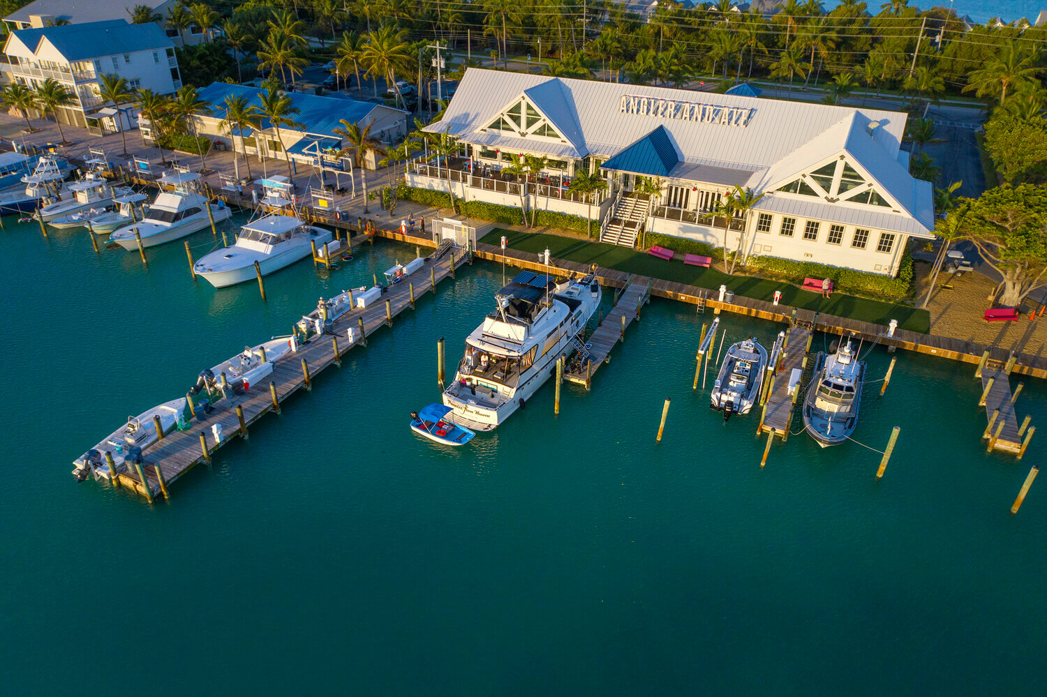 Aerial view of a waterfront marina with docked boats beside a coastal restaurant surrounded by palm trees.