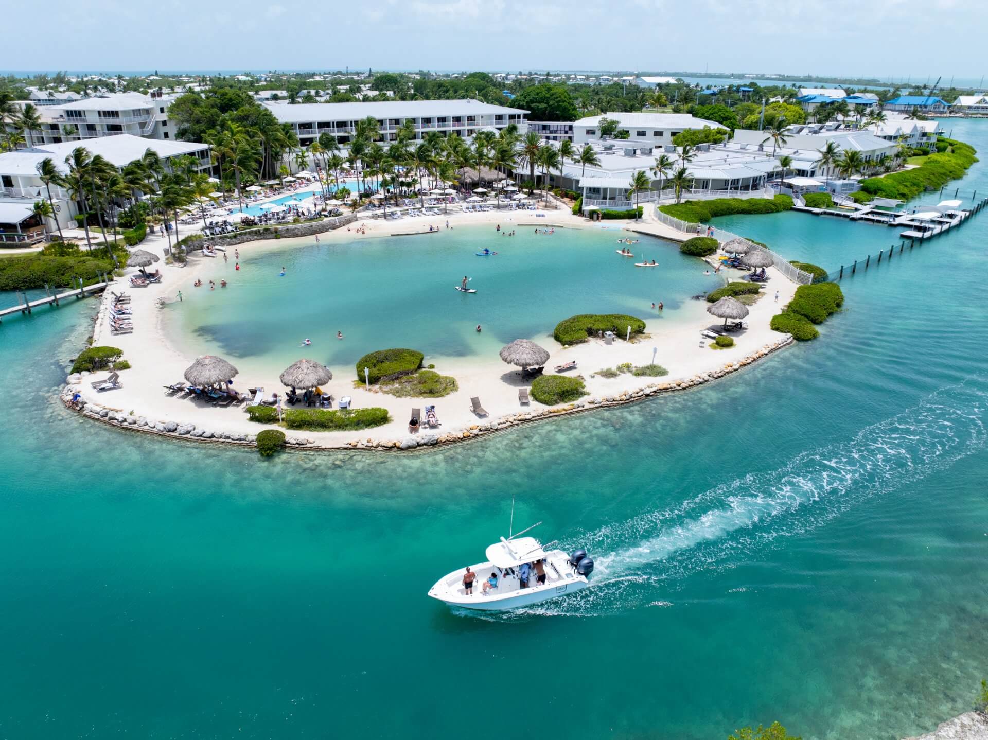 Aerial view of a tropical resort lagoon with sandy beach, palm-lined shoreline, and boats cruising turquoise waters.