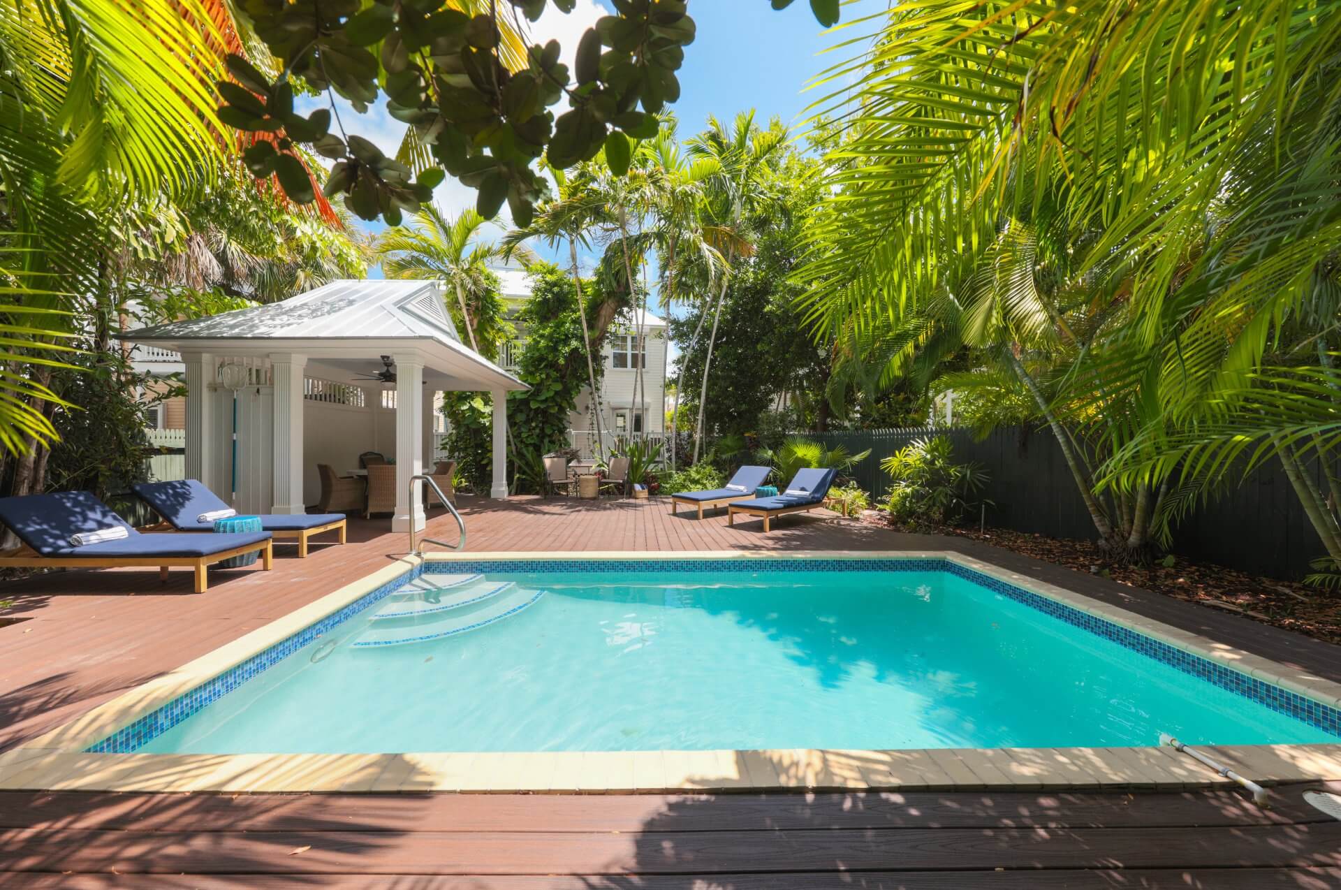 Tropical resort pool surrounded by palm trees, lounge chairs, and a shaded pavilion under a bright blue sky.